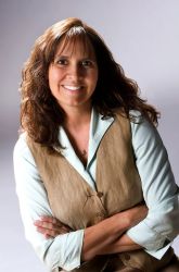 A smiling woman with shoulder-length brown hair wearing a light blue shirt and a beige vest sits with her arms crossed.